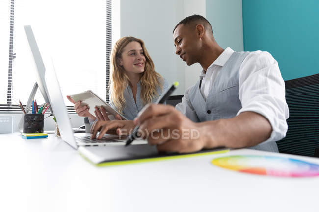 Seitenansicht eines jungen afrikanisch-amerikanischen Mannes, der einen Laptop und ein Grafik-Tablet benutzt, und einer jungen kaukasischen Frau, die an einem Schreibtisch sitzt und sich im modernen Büro eines kreativen Unternehmens unterhält — Stockfoto