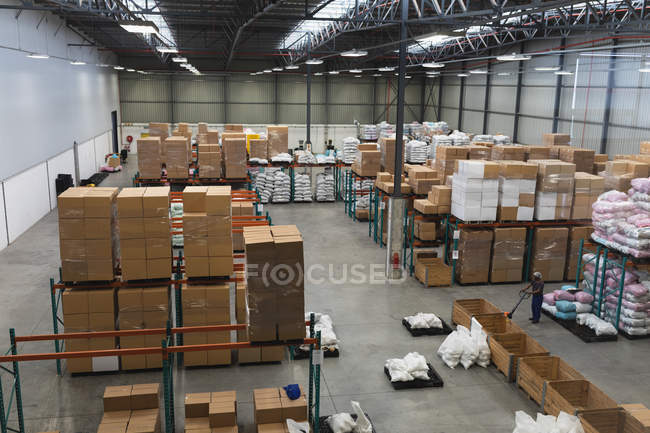High angle view of a young African American male factory worker using a trolley to move a crate in a product storage area in a warehouse at a factory — Stock Photo