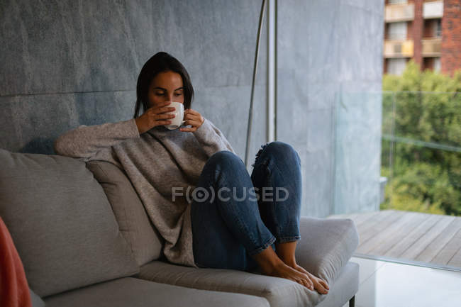 Vorderansicht einer jungen kaukasischen brünetten Frau, die mit hochgezogenen Beinen auf einem Sofa sitzt und eine Tasse Kaffee genießt — Stockfoto
