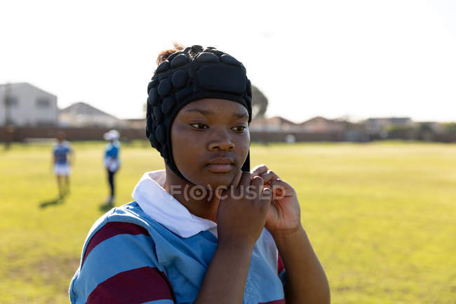 Vue de face gros plan d'une jeune joueuse de rugby mixte adulte debout sur un terrain de rugby attachant son protège-tête — Photo de stock