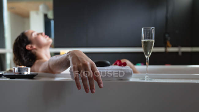 Side view of a young Caucasian brunette woman lying in a bath with a lit candle and a glass of champagne on the side, leaning back with her eyes closed — Stock Photo