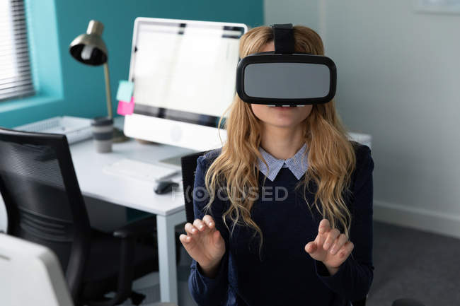 Вид спереди на молодую кавказскую женщину, сидящую в гарнитуре VR и с поднятыми руками в современном офисе творческого бизнеса, с пустым рабочим местом на заднем плане — стоковое фото