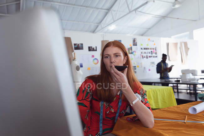 Vista frontale di una giovane studentessa caucasica di moda che tiene e parla su uno smartphone e usa un computer in uno studio all'università di moda, con uno studente che lavora sullo sfondo — Foto stock
