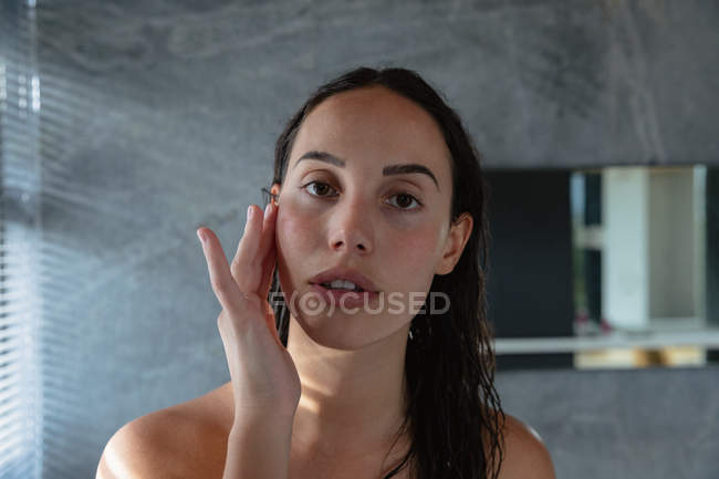 Porträt einer jungen kaukasischen brünetten Frau, die direkt in die Kamera blickt und ihr Gesicht in einem modernen Badezimmer mit einer Hand massiert — Stockfoto