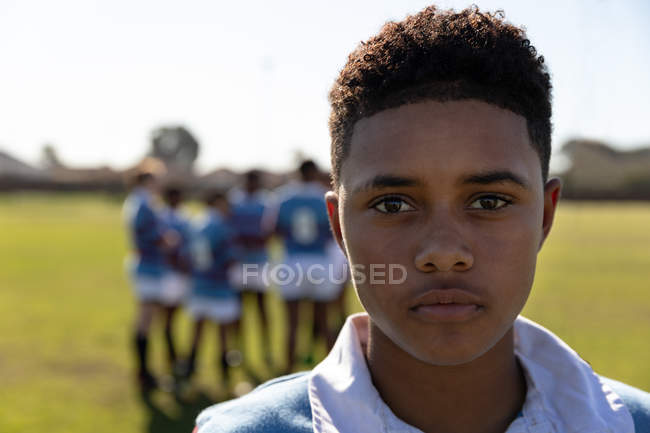 Portrait close up of a young adult mixed race female rugby player standing on a rugby pitch looking to camera, with her teammates talking together in the background — Stock Photo