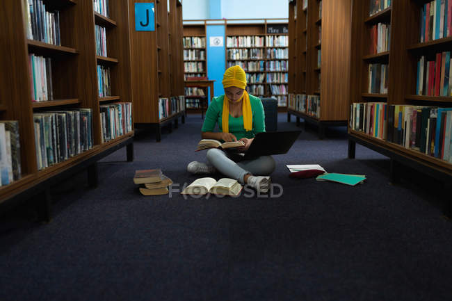 Front view of a young Asian female student wearing a hijab holding a book, using a laptop computer and studying in a library — Stock Photo