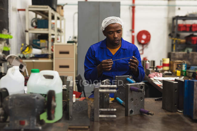 Front view close up of a young African American male factory worker sitting and inspecting equipment in the machine shop at a processing plant, with shelves of equipment in the background — Stock Photo