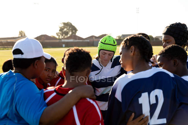 Visão traseira close-up de jovens adultos multi-étnicos jogadores de rugby do sexo feminino e sua meia idade mista treinador feminino em pé em um aconchego em um campo de rugby durante um jogo — Fotografia de Stock