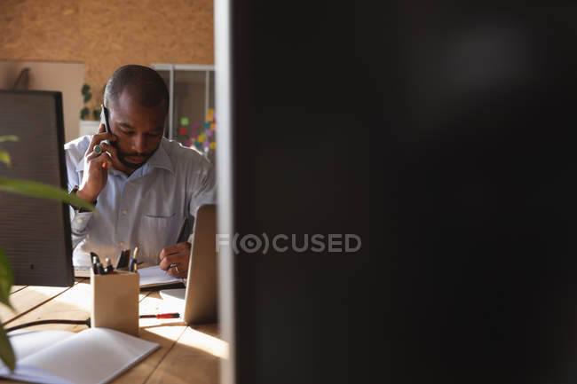 Nahaufnahme eines jungen afrikanisch-amerikanischen Mannes, der in einem kreativen Büro an einem Schreibtisch sitzt und mit einem Smartphone spricht, gesehen zwischen Computerbildschirmen — Stockfoto