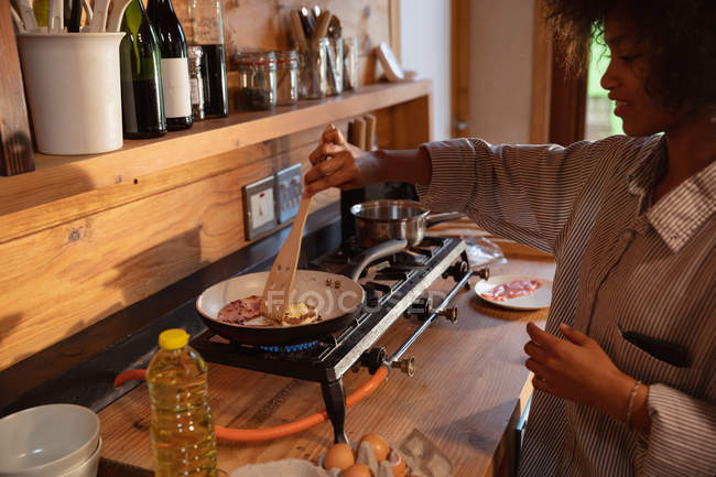 Seitenansicht einer lächelnden jungen Frau mit gemischter Rasse, die in ihrer Küche in einer Pfanne auf dem Herd das Frühstück zubereitet — Stockfoto