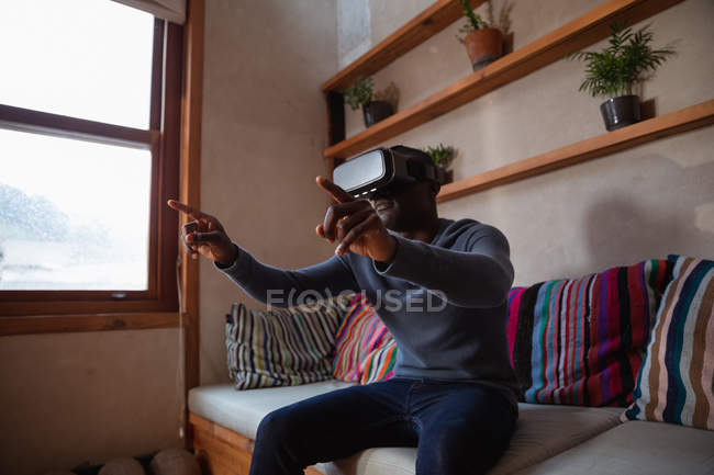 Side view close up of a young African American man wearing a VR headset sitting on a sofa at home, with his arms raised and fingers pointing — Stock Photo
