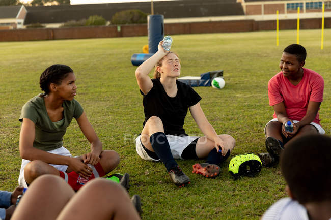 Frontansicht einer Mannschaft junger erwachsener multiethnischer Rugbyspielerinnen, die auf einem Rugbyfeld sitzen und sich während einer Trainingseinheit unterhalten und entspannen, wobei sich eine erfrischt, indem sie Wasser über ihren Kopf gießt. — Stockfoto