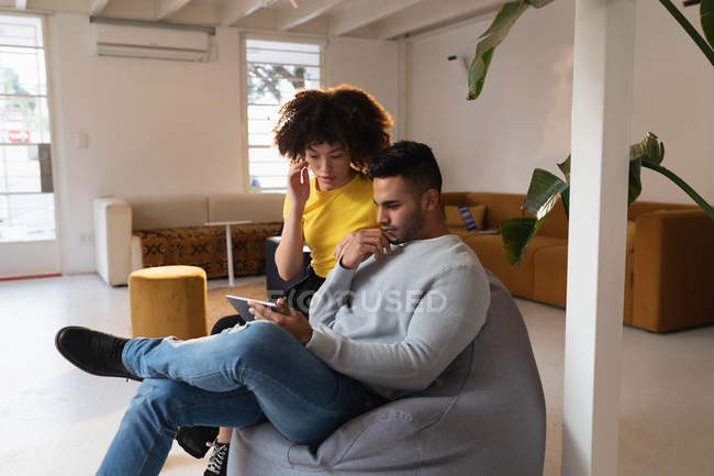 Вид спереду молодого змішаного чоловіка і молодої змішаної раси жінки, що сидить на мішку з бобами за допомогою планшетного комп'ютера і обговорює в творчому офісі — стокове фото