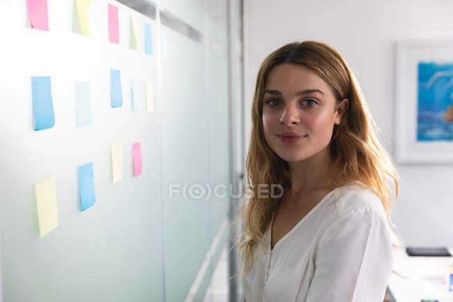 Портрет молодої кавказької жінки, що стоїть і посміхається до камери біля скляної стіни з кольоровими липкими нотатками на ній в сучасному офісі творчого бізнесу. — стокове фото