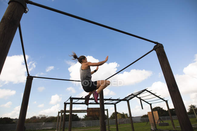 Vista lateral de una joven mujer caucásica en medio del aire balanceándose entre rejas en un marco en un gimnasio al aire libre durante una sesión de entrenamiento de bootcamp - foto de stock
