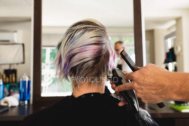 Vista posteriore primo piano di un parrucchiere caucasico di mezza età e di una giovane donna caucasica con i capelli tagliati in un parrucchiere, riflessi in uno specchio — Foto stock