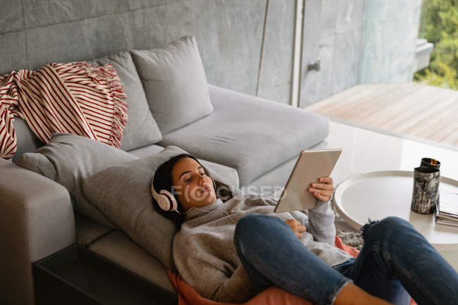 Elevated view of a young Caucasian brunette woman reclining on a sofa with her legs up, wearing headphones and watching a tablet computer — Stock Photo