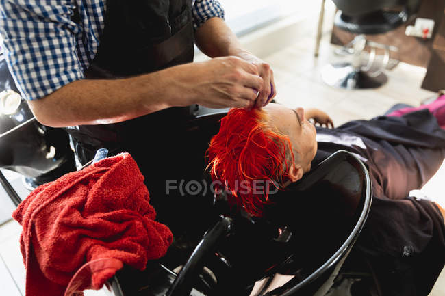 Vista laterale primo piano del parrucchiere maschile e di una giovane donna caucasica con i capelli colorati di rosso brillante e lavati in un salone di parrucchiere — Foto stock