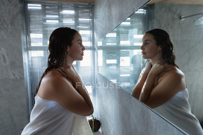Vista lateral de perto de uma jovem mulher morena caucasiana vestindo uma toalha de banho olhando no espelho e massageando o pescoço em um banheiro moderno — Fotografia de Stock