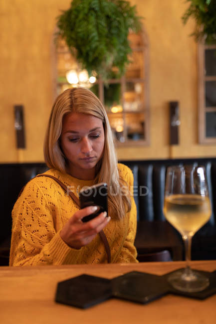 Vista frontal de una joven mujer caucásica relajándose de vacaciones en un bar, bebiendo vino y usando un teléfono inteligente - foto de stock