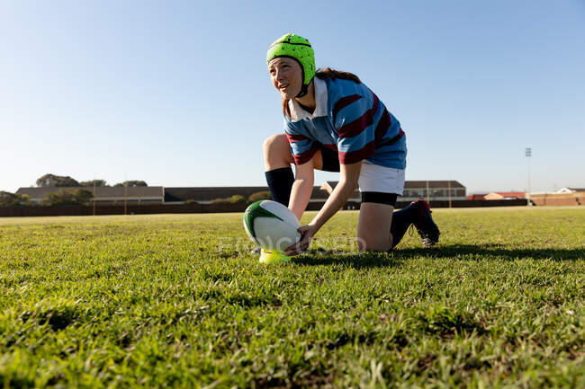 Vista frontal close up de um jovem adulto caucasiano jogador de rugby feminino vestindo um protetor de cabeça ajoelhado em um campo de rugby e definindo a bola em um tee para um chute de lugar — Fotografia de Stock