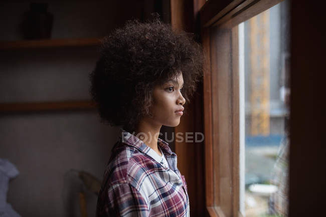 Vista laterale da vicino di una giovane donna di razza mista che indossa una camicia a quadri in piedi vicino a una finestra a casa guardando fuori — Foto stock