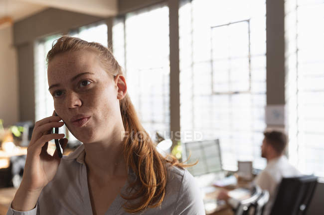 Вид спереди на молодую кавказку, разговаривающую по смартфону в креативном офисе, с коллегой, работающим на заднем плане — стоковое фото