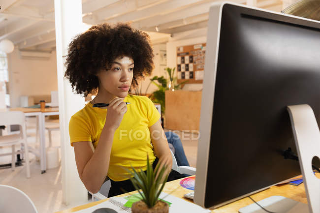 Передній вид крупним планом молода змішана жінка, що сидить за столом, дивлячись на екран комп'ютера в творчому офісі — стокове фото