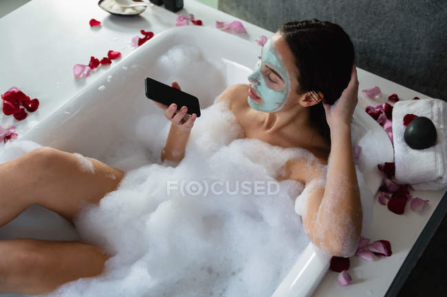 Высокий вид молодой белой женщины в упаковке для лица, сидящей в пенной ванне с лепестками роз по краю, используя смартфон — стоковое фото