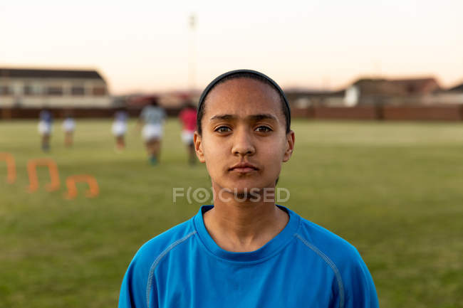 Portrait close up of a young adult mixed race female rugby player standing on a rugby pitch looking to camera, with her teammates in the background — Stock Photo