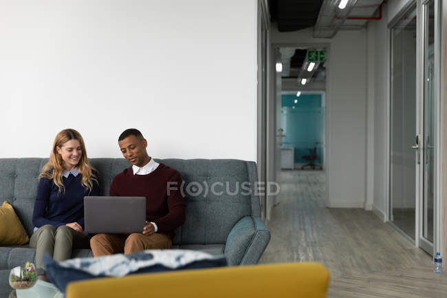 Vista frontal de um jovem afro-americano e uma jovem mulher caucasiana sentada olhando para um computador portátil e conversando sentada em um sofá na área de estar de um negócio criativo moderno — Fotografia de Stock
