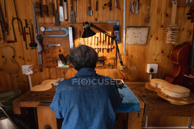 Rückansicht einer älteren kaukasischen Geigenbauerin, die in ihrer Werkstatt arbeitet, im Hintergrund hängen Werkzeuge an der Wand — Stockfoto