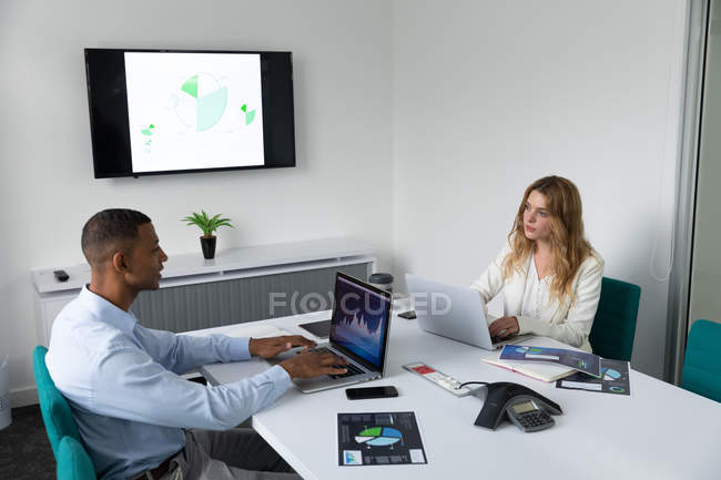 Vue latérale d'un jeune homme afro-américain et d'une jeune femme caucasienne assise sur les côtés opposés d'un bureau parlant et utilisant des ordinateurs portables dans le bureau moderne d'une entreprise créative — Photo de stock