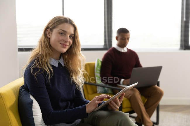 Вид сбоку на молодую кавказскую женщину с помощью планшетного компьютера и поворачивающуюся к камере улыбающуюся, сидящую в кресле в гостиной творческого бизнеса, с ее коллегой-мужчиной, сидящим на заднем плане с ноутбуком — стоковое фото