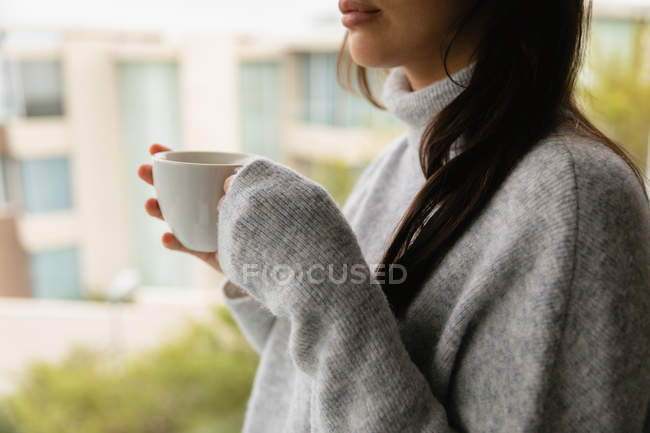 Вид збоку середині секції молодий Кавказький жінка брюнетка одягнений в сірий светр водолазки, стоячи на вікні, що тримає чашку кави — стокове фото