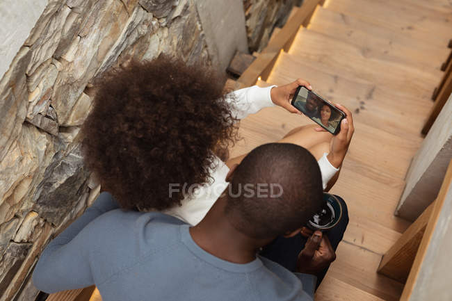 Vista posteriore sopraelevata di una giovane donna di razza mista e un giovane afroamericano che si fa un selfie con uno smartphone seduto sulle scale a casa . — Foto stock