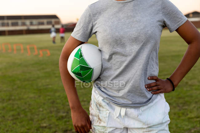 Передний вид средней части женского регбиста, стоящего на спортивном поле с рукой на бедре, держа регбийный мяч под мышкой во время тренировки — стоковое фото