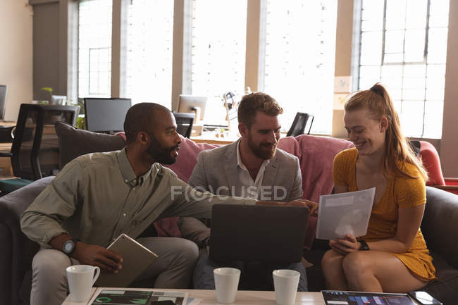 Front view close up of a young African American man and a young Caucasian woman and man sitting on a sofa in a creative office discussing a project and using a laptop and a tablet computer — Stock Photo
