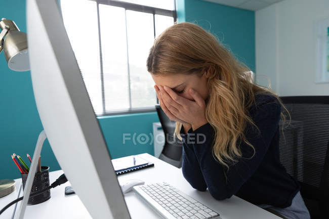 Вид сбоку на молодую кавказку, сидящую за столом за компьютером с головой в руках в современном офисе креативного бизнеса — стоковое фото