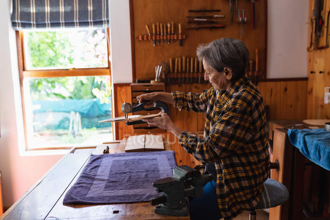 Вид збоку старшого Кавказького жіночого luthier вимірювання тіла скрипки в її майстерні з інструментами висить на стіні у фоновому режимі — стокове фото