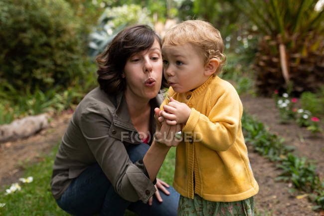 Nahaufnahme einer jungen kaukasischen Mutter, die mit ihrem Baby in einem Garten Löwenzahn bläst — Stockfoto