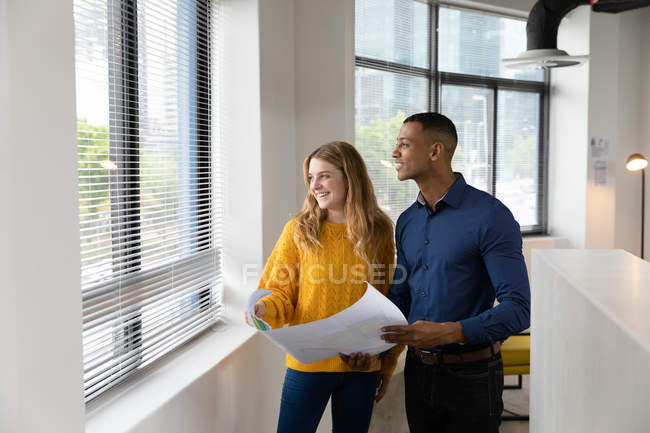 Vista frontal de un joven afroamericano y una joven mujer caucásica sosteniendo planes y mirando por la ventana juntos de pie en la oficina moderna de un negocio creativo - foto de stock