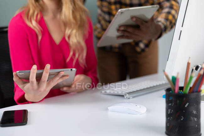 Vue de face au milieu d'un jeune homme afro-américain debout et d'une jeune femme caucasienne assise à un bureau à l'aide d'ordinateurs tablettes dans le bureau moderne d'une entreprise créative — Photo de stock