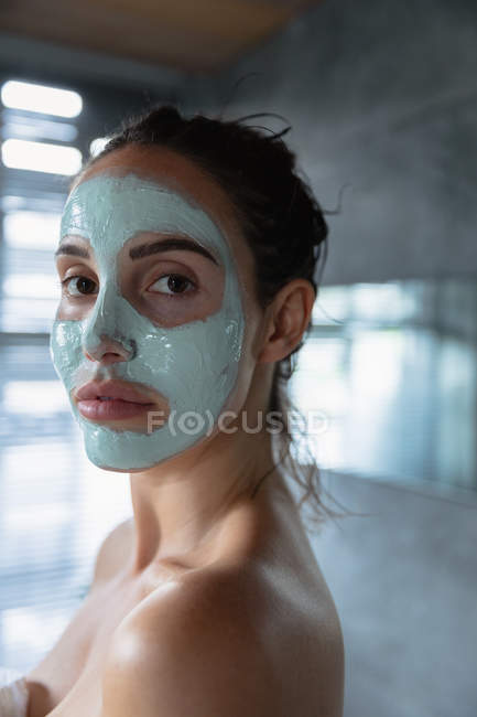 Vista lateral de cerca de una joven morena caucásica que lleva un paquete facial girando para mirar a la cámara en un baño moderno - foto de stock