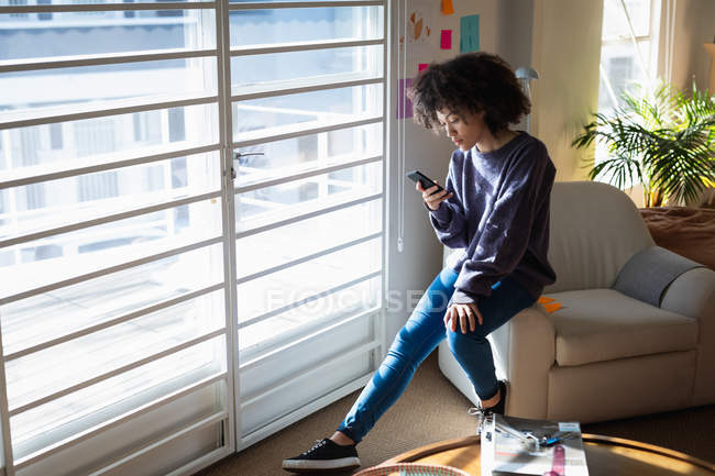 Vista lateral de uma jovem mulher de raça mista sentada em uma poltrona e usando um smartphone em um escritório criativo — Fotografia de Stock