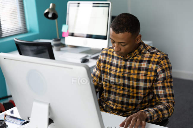 Vista frontale di un giovane afroamericano seduto ad una scrivania con un computer nell'ufficio moderno di un'azienda creativa, con una postazione di lavoro vuota sullo sfondo — Foto stock