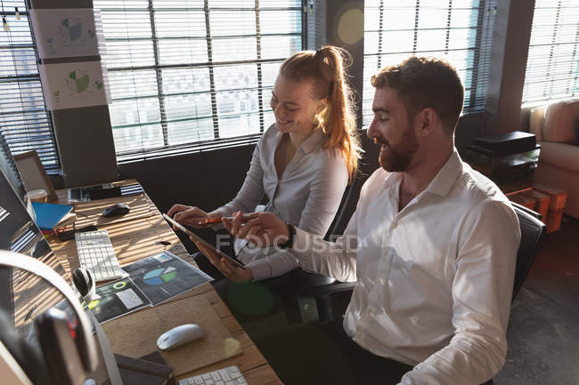 Vista lateral elevada de una joven mujer y un hombre caucásicos sentados en un escritorio sonriendo mirando una tableta en una oficina creativa - foto de stock
