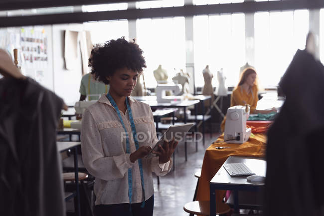 Vista frontal de una joven estudiante de moda de raza mixta usando una tableta mientras trabaja en un diseño en un estudio en la universidad de moda, con otros estudiantes en el trabajo en segundo plano - foto de stock