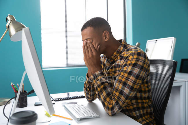 Вид сбоку на молодого афроамериканца, сидящего за столом за компьютером с головой в руках в современном офисе креативного бизнеса — стоковое фото