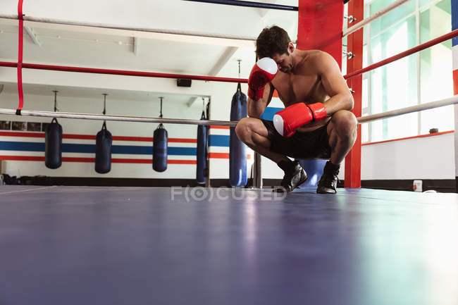 Vista frontal perto de um jovem boxeador caucasiano agachado em um anel de boxe descansando a cabeça em uma luva de boxe — Fotografia de Stock
